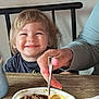 child, smiling, blond_hair, happy, fork, food, macaroni, meat, table, plate, hand, indoor, meal, person, toddler, hair, face, wooden_table, casual_clothing, dining