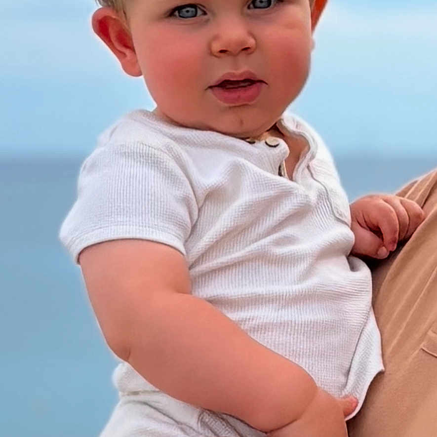 Eden a rejoint le concours — aidez-le/la à gagner de superbes lots ! adult_hand, baby, blonde_hair, blue_eyes, casual_clothing, child, cute, daylight, expression, face, hand, holding, ocean, outdoor, person, portrait, sky, summer, toddler, white_shirt