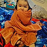 baby, towel, blanket, foot, face, child, indoors, pillow, toy, window, curly_hair, colorful, bed, wrapped, sitting, cute, looking, home, soft, comfort
