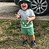 toddler, child, smiling, laughing, outdoor, grass, dinosaur_hat, dinosaur_shirt, green_clothing, shorts, sneakers, car_wheel, driveway, happy, cute, person, young_child, nature, playful, summer_clothes