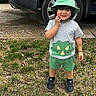 toddler, child, smiling, outdoor, grass, car, hat, shorts, dinosaur, green_clothing, gray_shirt, black_shoes, sidewalk, young_child, cute, playful, casual, portrait, person, nature