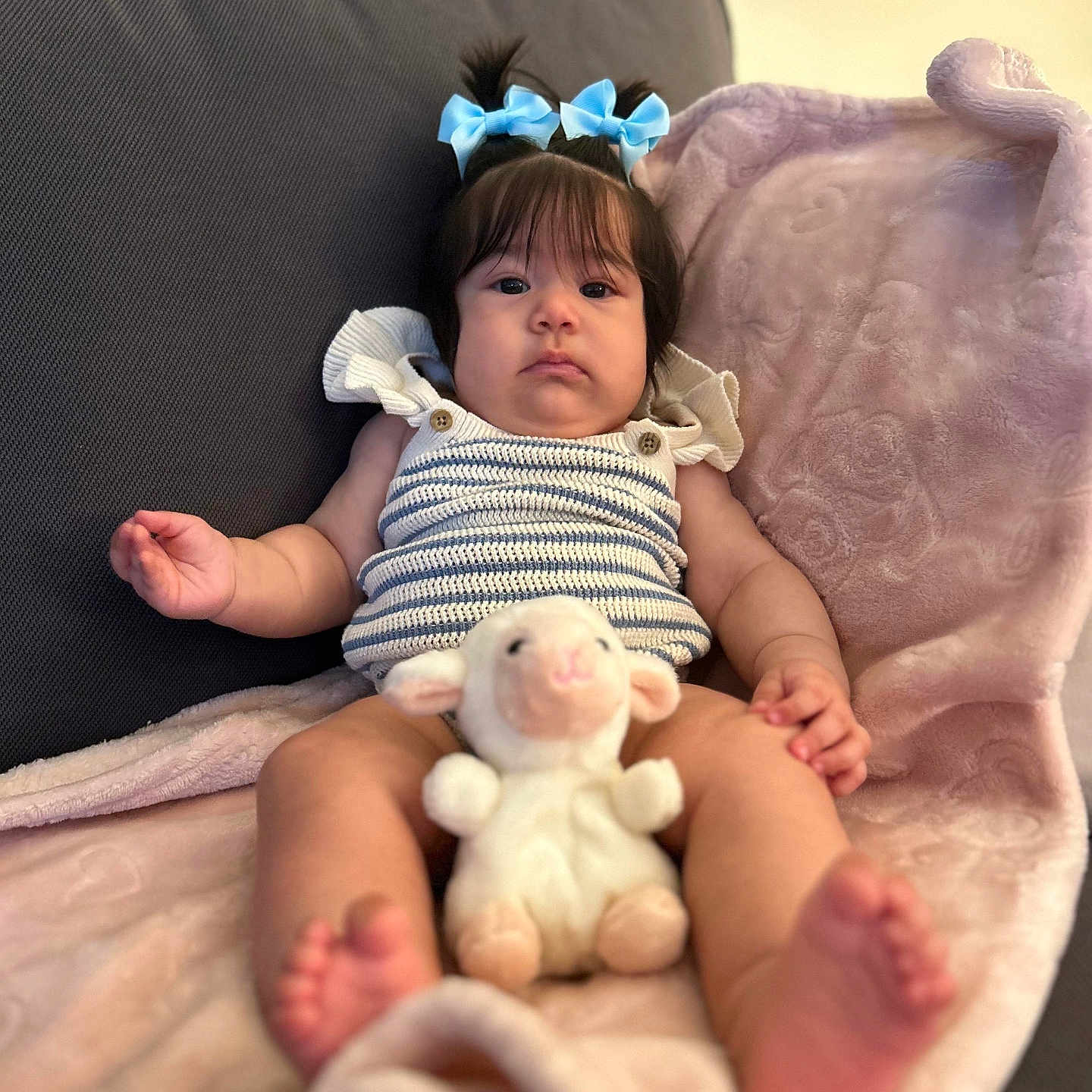 Akemy is registered to the contest to win money with this photo: baby, child, blue_bows, plush_toy, lamb, blanket, couch, striped_clothing, indoor, soft_toy, infant, cute, portrait, person, relaxing, seated, pink, furniture, hair_bow, expression