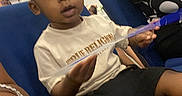 Allandrius is registered to the contest to win money with this photo: toddler, child, person, blue_chair, white_shoe, black_shorts, light_shirt, hairstyle, indoor, seated, holding_object, patterned_clothing, young_child, casual_clothing, curious_expression, people, furniture, footwear, portrait, event