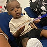toddler, child, person, blue_chair, white_shoe, black_shorts, light_shirt, hairstyle, indoor, seated, holding_object, patterned_clothing, young_child, casual_clothing, curious_expression, people, furniture, footwear, portrait, event