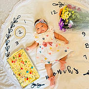 Mikaela is registered to the contest to win money with this photo: baby, infant, girl, dress, headband, flowers, bouquet, cake, sprinkles, candy, blanket, numbers, embroidery, pink_socks, celebration, milestone, smile, lying_down, cute, indoors