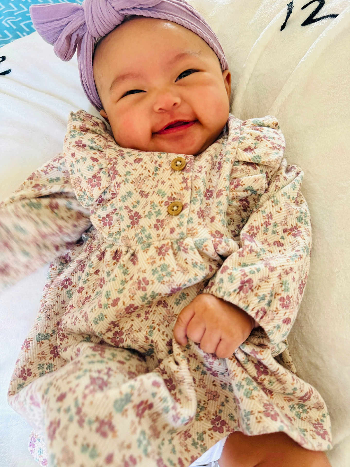 Mikaela joined the competition — help win amazing prizes! baby, smiling, headband, bow, floral_dress, infant, cute, happy, hand, clothing, portrait, child, lying_down, soft_texture, white_background, button, fabric, skin, cheeks, joy