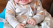 Teegan is registered to the contest to win money with this photo: baby, child, infant, smiling, happy, portrait, sitting, onesie, blanket, couch, home, indoor, wooden_floor, brown_hair, face, hands, feet, patterned_clothing, family, cozy