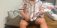 Ezekiah is registered to the contest to win money with this photo: toddler, child, smiling, indoor, table, baseball_jersey, mickey_mouse, shorts, sneakers, black_shoes, necklace, wooden_table, clothing, happy, person, diapers, bags, casual, background, furniture