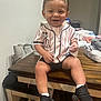 toddler, child, smiling, indoor, table, baseball_jersey, mickey_mouse, shorts, sneakers, black_shoes, necklace, wooden_table, clothing, happy, person, diapers, bags, casual, background, furniture