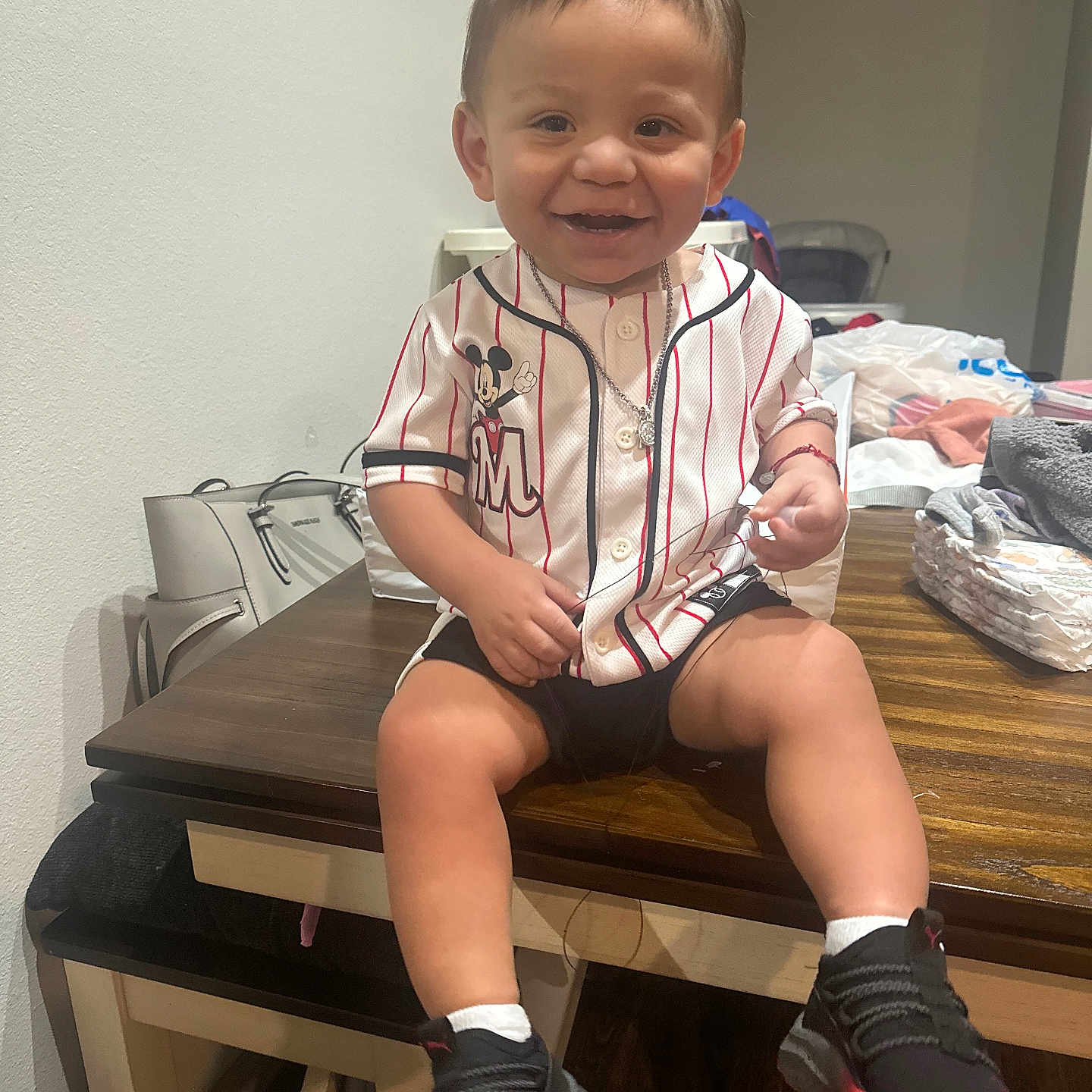 Ezekiah is registered to the contest to win money with this photo: background, bags, baseball_jersey, black_shoes, casual, child, clothing, diapers, furniture, happy, indoor, mickey_mouse, necklace, person, shorts, smiling, sneakers, table, toddler, wooden_table
