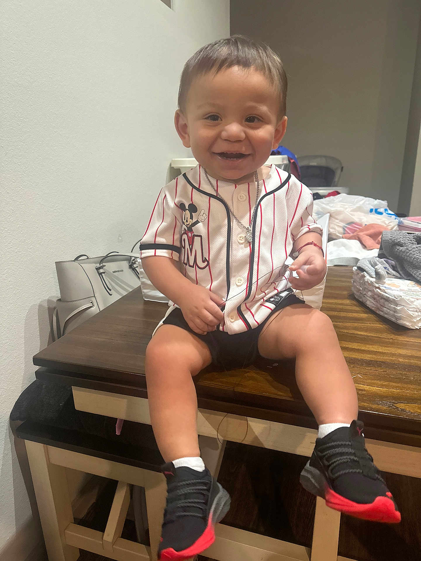 Ezekiah is registered to the contest to win money with this photo: toddler, child, smiling, indoor, table, baseball_jersey, mickey_mouse, shorts, sneakers, black_shoes, necklace, wooden_table, clothing, happy, person, diapers, bags, casual, background, furniture
