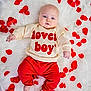 baby, blue_eyes, boy, child, clothing, cream_sweatshirt, cute, face, happy, indoor, infant, lying_down, portrait, red_pants, red_text, rose_petals, skin, smiling, soft_texture, white_rug