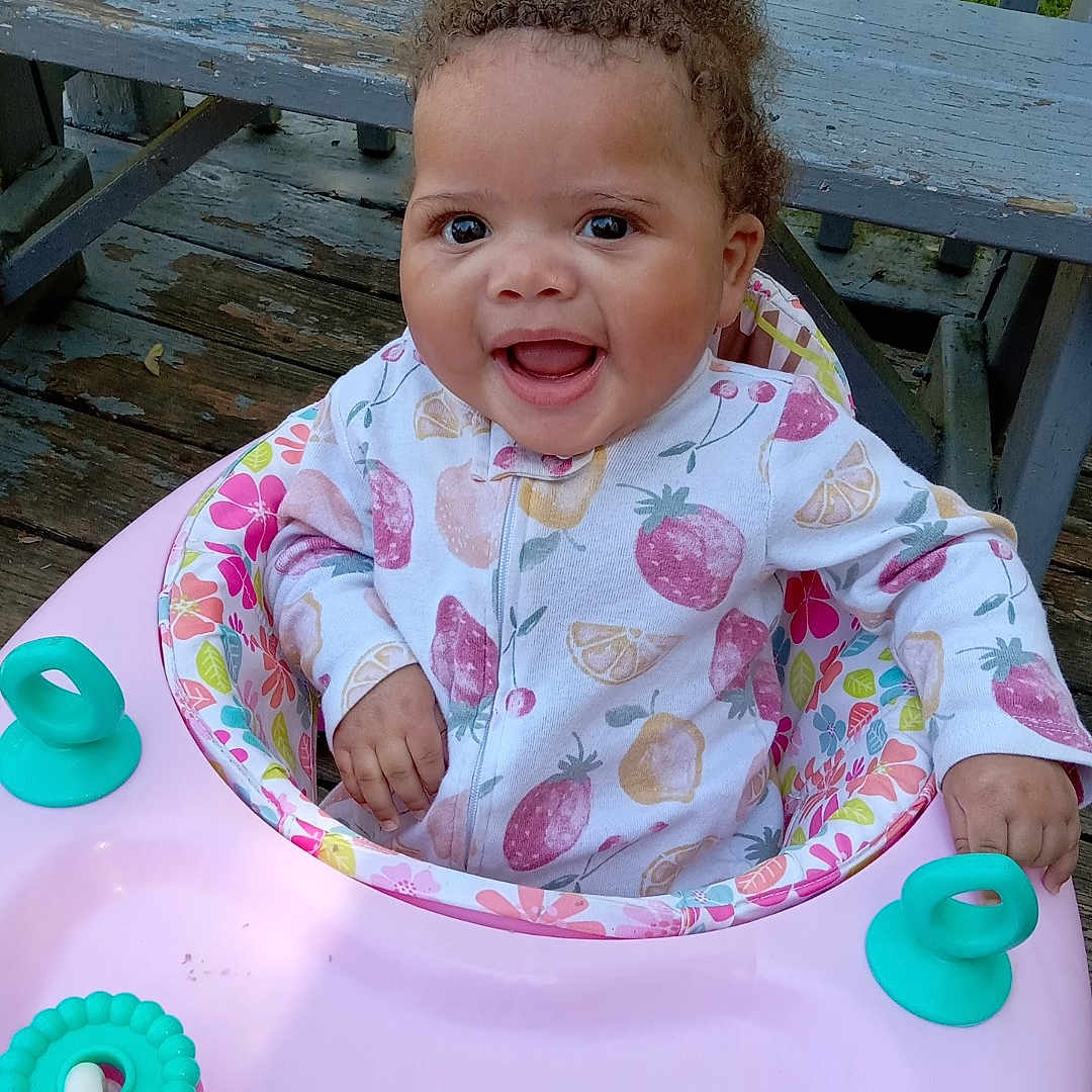 Tyleigh joined the competition — help win amazing prizes! baby, child, colorful_clothing, curly_hair, cute, grass, happy, infant, onesie, orange, outdoor, person, pink, playful, seat, smiling, teal, toy, walker, wooden_deck