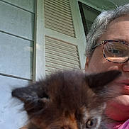 Flower is registered to the contest to win money with this photo: person, glasses, kitten, cat, close_up, hand, window, wall, shirt, hair, face, outdoor, portrait, animal, pet, holding, blur, eyewear, skin, home