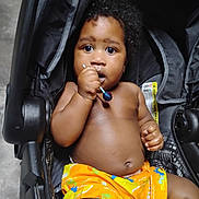 Ace joined the competition — help win amazing prizes! baby, child, stroller, lollipop, orange_shorts, curly_hair, dark_skin, sitting, indoors, close_up, cute, expression, hand, face, infant, person, young_child, bright_colors, looking, toy