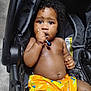 baby, child, stroller, lollipop, orange_shorts, curly_hair, dark_skin, sitting, indoors, close_up, cute, expression, hand, face, infant, person, young_child, bright_colors, looking, toy