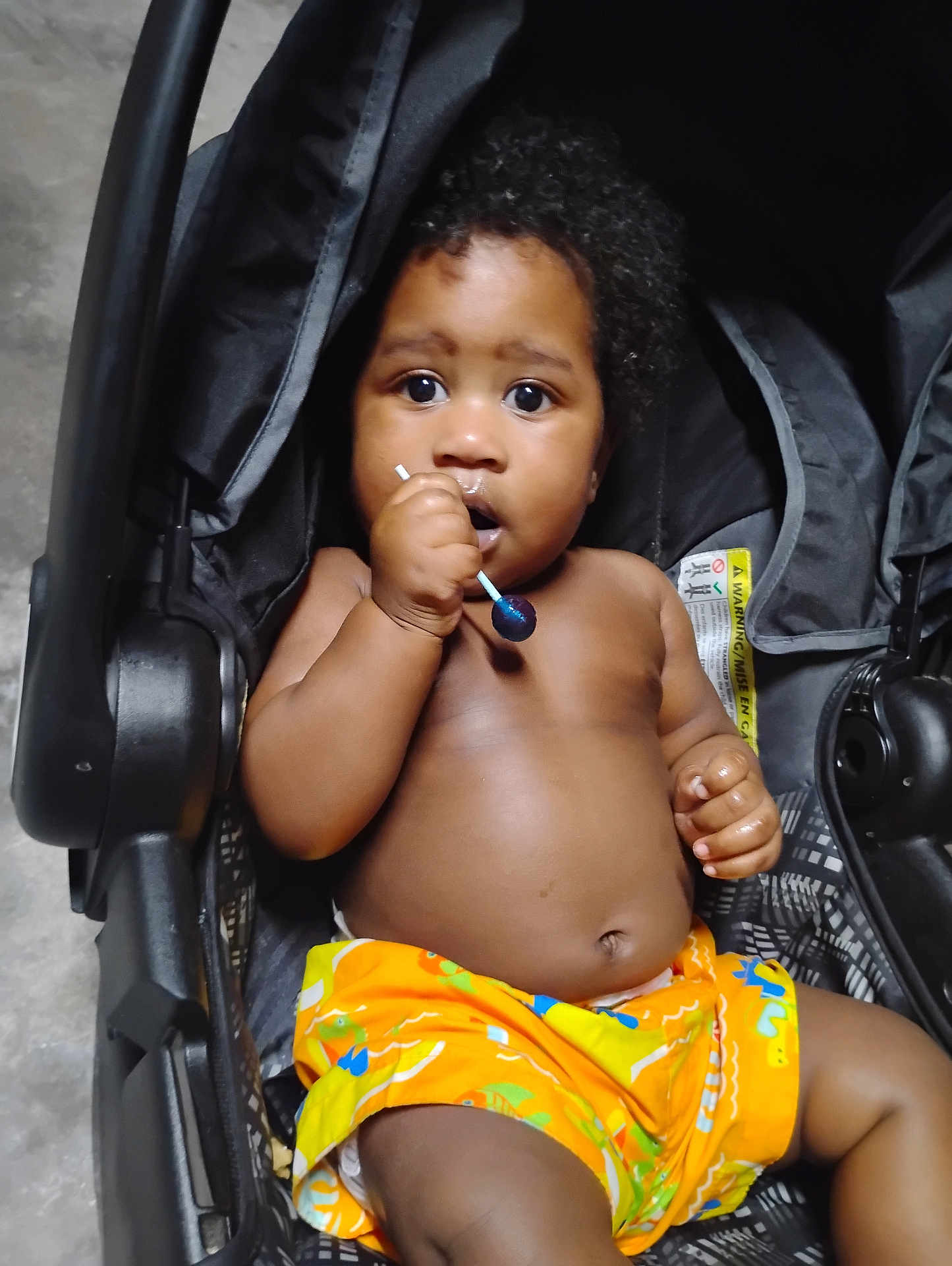 Ace joined the competition — help win amazing prizes! baby, child, stroller, lollipop, orange_shorts, curly_hair, dark_skin, sitting, indoors, close_up, cute, expression, hand, face, infant, person, young_child, bright_colors, looking, toy