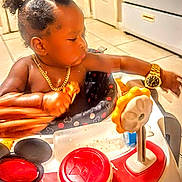 Ace is registered to the contest to win money with this photo: baby, black_hair, child, curly_hair, cute, door, floor_tiles, food, gold_chain, indoors, infant, kitchen, person, plastic_toy, puffs_hairstyle, red_button, toy, walker, watch, yellow_gear
