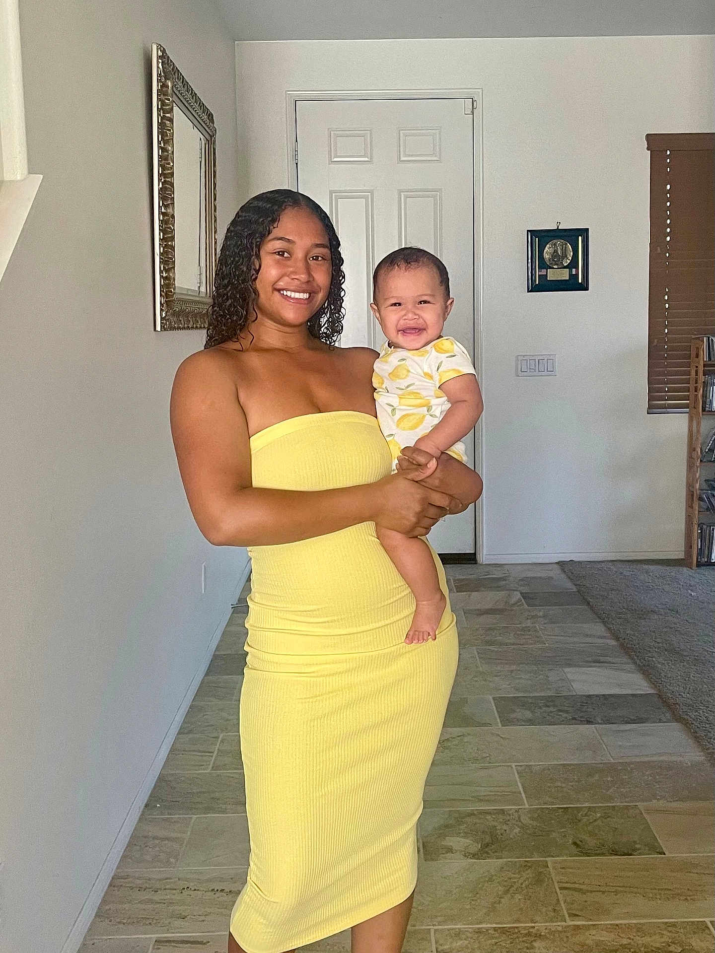 Lehlani joined the competition — help win amazing prizes! woman, baby, smile, yellow_dress, indoor, tile_floor, mirror, door, holding, child, happy, person, curly_hair, short_sleeves, plaid, home_interior, window_blinds, carpet, shelf, photography