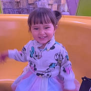 Leilanny is registered to the contest to win money with this photo: child, girl, smiling, jacket, tutu_skirt, shoes, yellow_seat, amusement_ride, rock_background, indoor, playful, happy, person, casual_clothing, short_hair, colorful, portrait, fun, cute, young