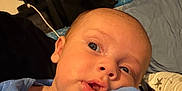 Ryker is registered to the contest to win money with this photo: baby, infant, closeup, blue_clothing, hand, face, eyes, indoors, bed, blanket, expression, person, newborn, skin, portrait, sleepwear, cute, young_child, resting, curious