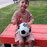Bryson is registered to the contest to win money with this photo: child, boy, soccer_ball, bench, outdoor, grass, tree, building, spiderman, shorts, tshirt, footwear, blue_slippers, park, play, smile, daylight, casual_clothing, recreation, young