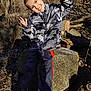 Bryson is registered to the contest to win money with this photo: child, smiling, outdoor, forest, rock, camouflage_jacket, pants, shoes, sunlight, trees, nature, playful, pose, happy, young, person, casual_clothing, daylight, ground, leaves