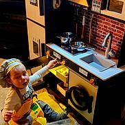 Ian joined the competition — help win amazing prizes! toddler, child, toy_kitchen, apron, chef_hat, play, indoor, carpet, pot, pan, pretend_play, plastic_container, knobs, sink, stove, oven, cabinet, faucet, cooking, toy