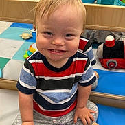 Ari is registered to the contest to win money with this photo: child, baby, toddler, smiling, happy, portrait, sitting, toy_store, toys, rattles, blocks, plush_toy, colorful, striped_shirt, gray_pants, socks, play_area, indoor, wooden_shelf, face