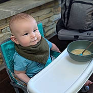 Wyatt is registered to the contest to win money with this photo: baby, high_chair, bib, blue_shirt, bowl, spoon, food, feeding, infant, smiling, face, cheeks, furniture, stone_wall, backpack, indoor, floor, wooden_floor, person, child