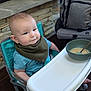 baby, high_chair, bib, blue_shirt, bowl, spoon, food, feeding, infant, smiling, face, cheeks, furniture, stone_wall, backpack, indoor, floor, wooden_floor, person, child