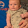 baby, child, infant, person, smile, arm, hand, clothing, onesie, pattern, tree_pattern, indoor, furniture, crib, skin, face, eyes, cheeks, person_holding, comfort