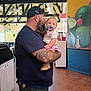 Emma Pauline is registered to the contest to win money with this photo: man, baby, child, tattoo, pacifier, cap, indoor, restaurant, exit_sign, flooring, wall_art, beard, holding, clothing, person, shoes, table, chair, lighting, window