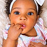 baby, blanket, bow, child, close_up, colorful_clothing, curly_hair, face, floral_dress, fluffy_blanket, hand, headband, indoor, infant, lying_down, portrait, self_soothing, sitting, smiling, wide_eyes