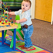 Theo is registered to the contest to win money with this photo: toddler, child, indoor, playroom, toy, bead_maze, colorful, carpet, shirt, jeans, brown_shoes, curly_hair, table, chair, wooden_toy, educational, learning, young_child, standing, curious