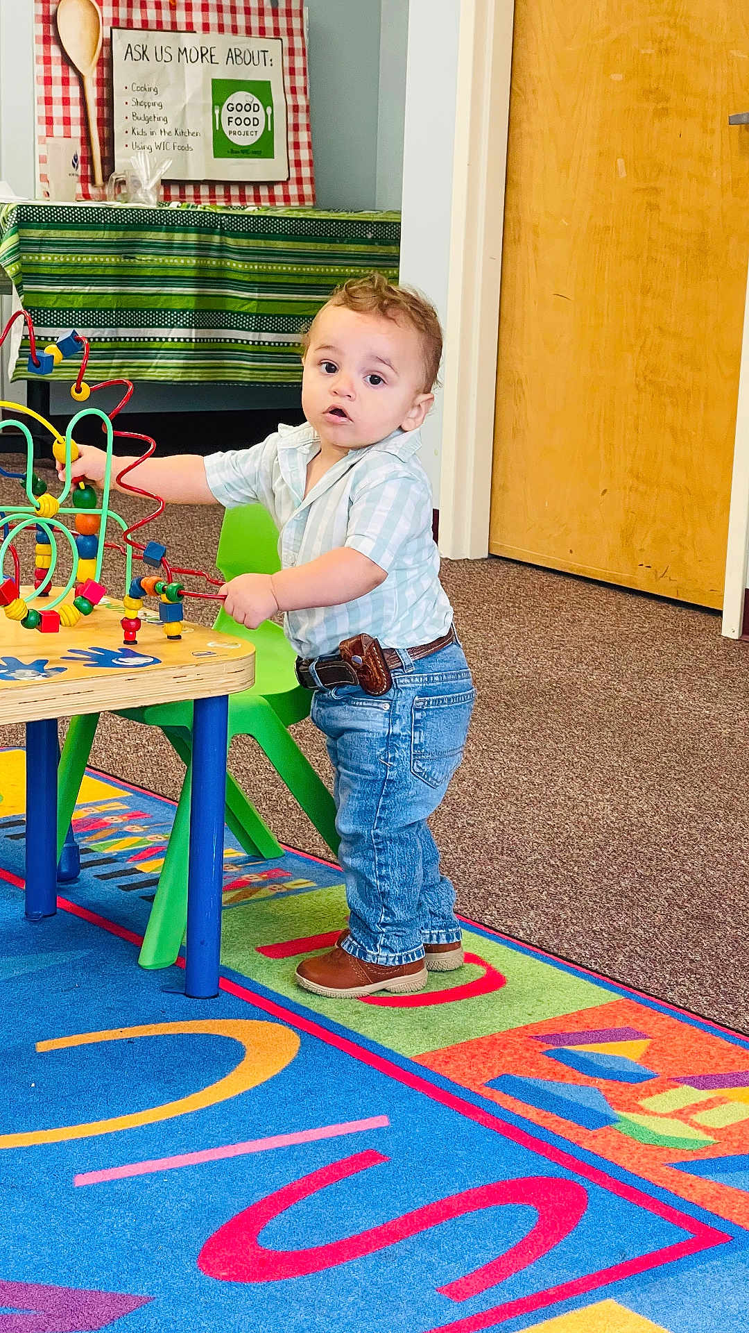 Theo is registered to the contest to win money with this photo: toddler, child, indoor, playroom, toy, bead_maze, colorful, carpet, shirt, jeans, brown_shoes, curly_hair, table, chair, wooden_toy, educational, learning, young_child, standing, curious