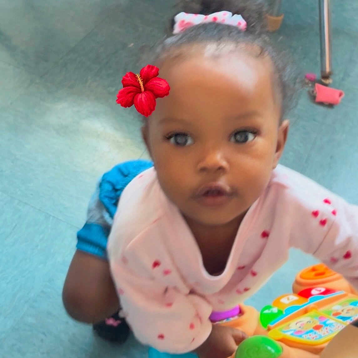 Jasamira is registered to the contest to win money with this photo: blue_shorts, candid, child, colorful, curly_hair, cute, emotion, expression, floor, flower_hair_accessory, furniture_legs, indoor, looking_up, pink_sweater, plastic_toy, playroom, playtime, toddler, toy, young_child