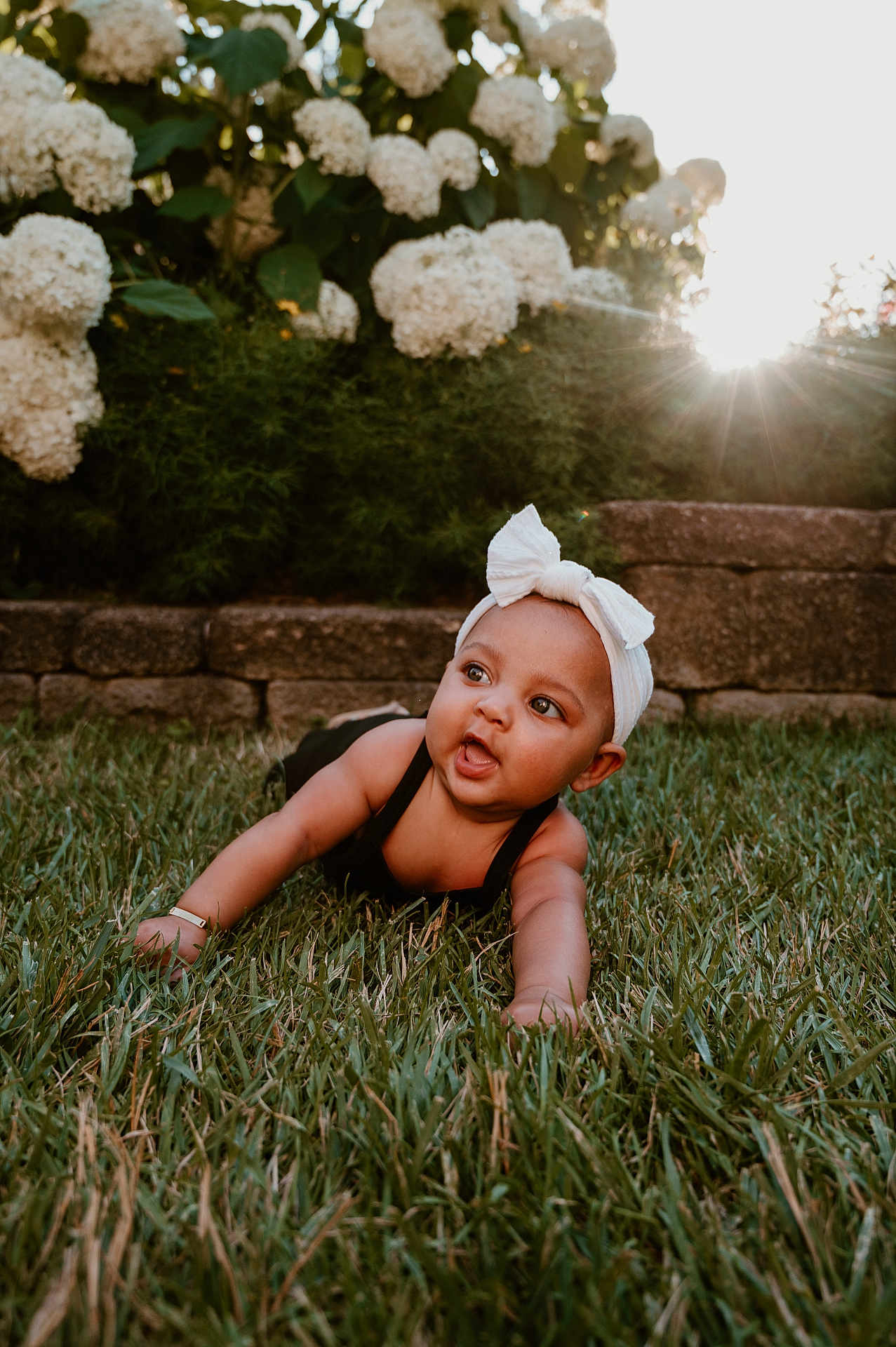 Tala joined the competition — help win amazing prizes! baby, child, grass, flower, garden, outdoor, headband, bow, sunlight, stone_wall, crawling, greenery, nature, cute, young_child, portrait, daylight, happy, face, skin