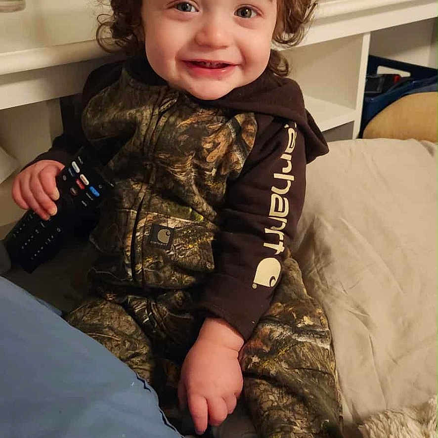 Yz Hezekiah joined the competition — help win amazing prizes! bed, blanket, camo_clothing, child, cozy, curly_hair, curtain, face, furniture, happy, home, indoor, person, pillow, plant, remote_control, sitting, smiling, sock, toddler