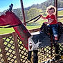 toddler, child, toy_horse, wooden_horse, red_shirt, mickey_mouse, jeans, barefoot, curly_hair, outdoor, sunlight, fence, greenery, grass, trees, rural, play, sitting, holding_reins, daytime