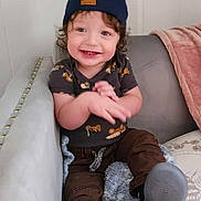 Yz Hezekiah is registered to the contest to win money with this photo: toddler, child, smiling, happy, sitting, beanie, curly_hair, blanket, couch, clothing, footwear, brown_pants, blue, indoors, person, cute, portrait, baby, hands, expression
