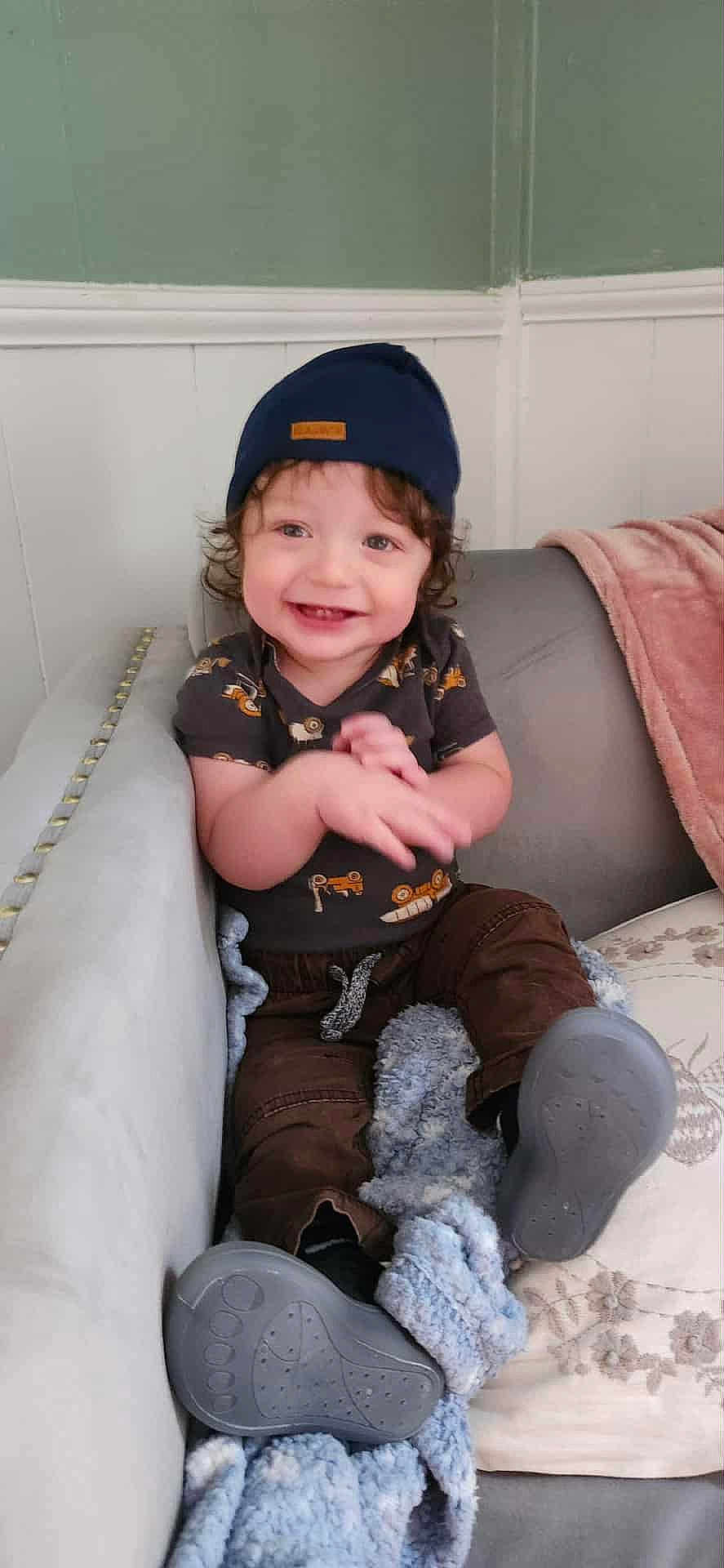 Yz Hezekiah is registered to the contest to win money with this photo: toddler, child, smiling, happy, sitting, beanie, curly_hair, blanket, couch, clothing, footwear, brown_pants, blue, indoors, person, cute, portrait, baby, hands, expression