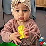 toddler, child, building_block, toy, headwrap, knit_sweater, indoor, floor, green_container, colorful, blue_eyes, hands, living_room, wooden_cabinet, television, playtime, curious, plastic_toy, floor_tiles, furniture