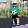 Aarron-Junior is registered to the contest to win money with this photo: blacktop, child, dinosaur_hat, fence, focused, grass, green_jacket, hat, outdoor, person, playground, purple_surface, shadow, shorts, sneakers, soccer_ball, sportswear, sunlight, toddler, young_child