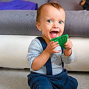 Tiago participe au concours pour gagner de l'argent avec cette photo : baby, child, toy, green_toy, smiling, happy, indoor, floor, sitting, clothing, striped_shirt, suspenders, pants, face, person, furniture, sofa, cushion, home, play