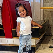 Zoe is registered to the contest to win money with this photo: toddler, child, smile, standing, jeans, sneakers, white_shirt, hair_tie, indoor, tile_floor, booth, red_cushion, wall, person, happy, casual_clothing, young_child, footwear, cute, portrait