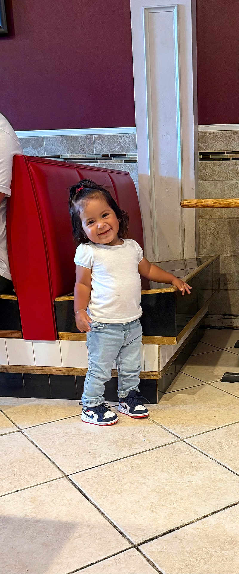 Zoe is registered to the contest to win money with this photo: toddler, child, smile, standing, jeans, sneakers, white_shirt, hair_tie, indoor, tile_floor, booth, red_cushion, wall, person, happy, casual_clothing, young_child, footwear, cute, portrait