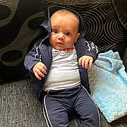 Lorenzo is registered to the contest to win money with this photo: baby, child, infant, hoodie, tracksuit, couch, blanket, pillow, curious, seated, clothing, indoor, portrait, face, hand, expression, fabric, text_on_clothing, blue, white
