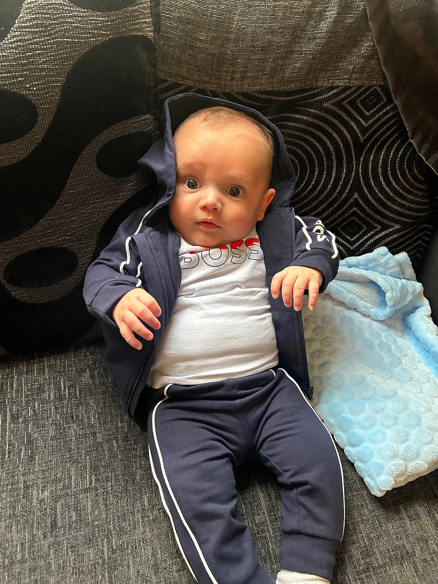 Lorenzo is registered to the contest to win money with this photo: baby, child, infant, hoodie, tracksuit, couch, blanket, pillow, curious, seated, clothing, indoor, portrait, face, hand, expression, fabric, text_on_clothing, blue, white