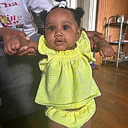 Aziaha joined the competition — help win amazing prizes! baby, child, infant, yellow_outfit, hair_bow, indoor, wooden_floor, holding_hands, curious_expression, person, adult_hands, bright_clothing, standing, home_interior, flooring, furniture, portrait, cute, small_child, people